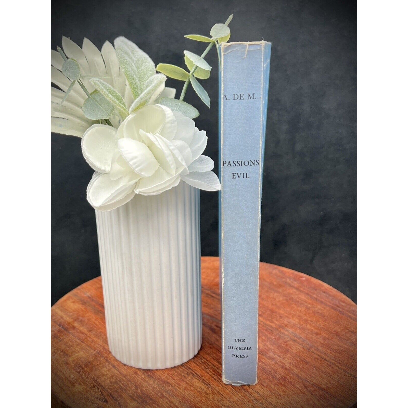 Blue Olympia Press 1953 Passions Evil by Alfred de Musset, numbered copy on a wooden table beside white flowers.
